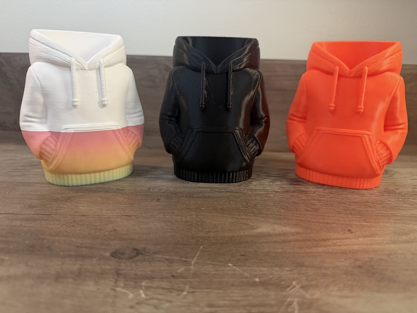 Three hoodie-shaped desk cups in white-to-orange gradient, black, and orange